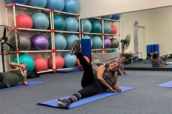 Moorundie Ruwe Ninkawi's Group fitness session Three women are lying on mats on the ground, laughing with their legs in the air. There is a shelf of exercise balls behind them