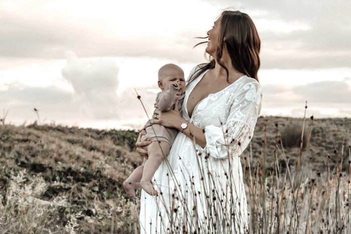 Danielle Ford with her daughter, Sienna Ford A mother in a loose white dress with long brown hair holds a baby wearing a white romper in a field.