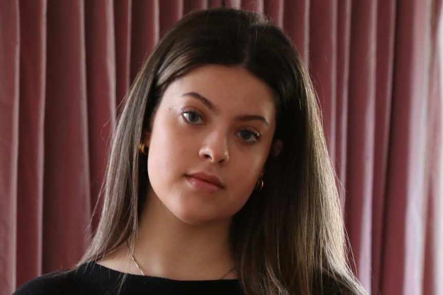 A young woman stands in a living room holding school books.