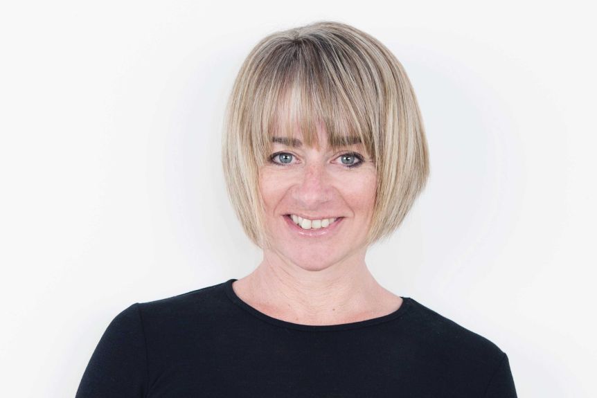 A woman with short blond hair and black top stands in front of a white background.