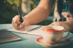 a woman sitting at a table: A journal is really just a notebook that can have any number of functions. You can use it as a calendar or daily planner, or pour your darkest secrets into it. Let’s take a look at some of the best ways to use a journal and how they can benefit us. Click through the gallery to get started.